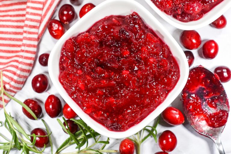 a bowl of bright red cranberry compote surrounded by cranberries and rosemary