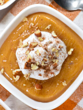 A bowl of butternut squash pudding topped with coconut whipped cream, on an orange checkered towel with a spoon