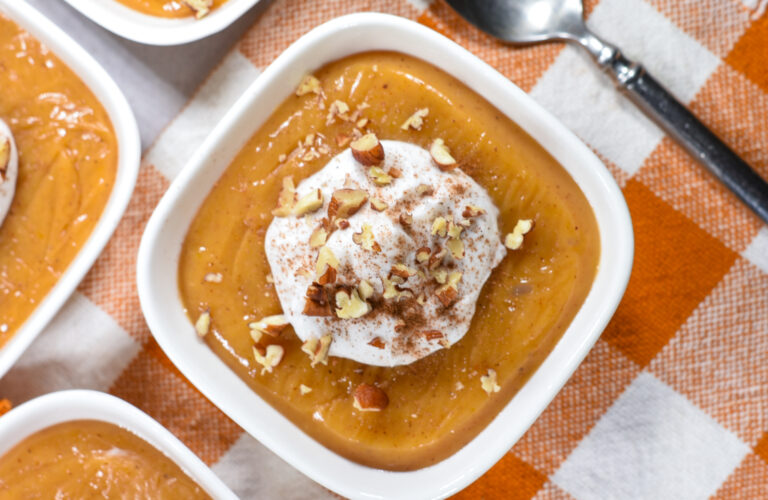 A bowl of butternut squash pudding topped with coconut whipped cream, on an orange checkered towel with a spoon