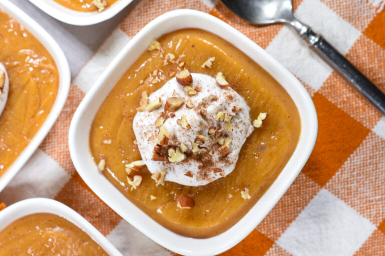 A bowl of butternut squash pudding topped with coconut whipped cream, on an orange checkered towel with a spoon