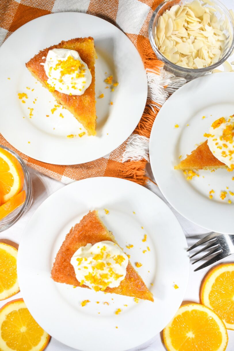 Flourless orange almond cake slices on white plates, with a checkered tea towel and fresh orange slices