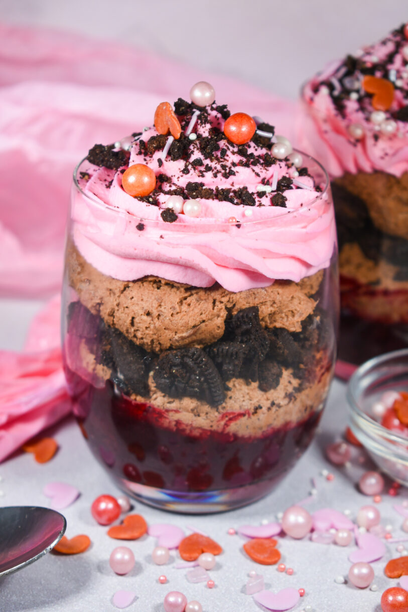 A Valentine parfait in a clear glass showing layers of cherry compote, chocolate mousse, Oreos and pink whipped cream