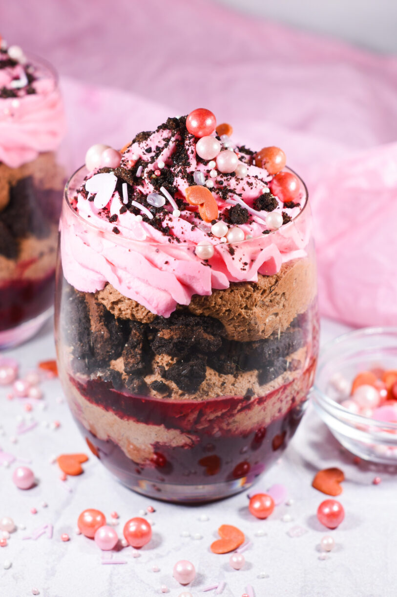 A parfait in a clear glass showing layers of cherry compote, chocolate mousse, Oreos and pink whipped cream