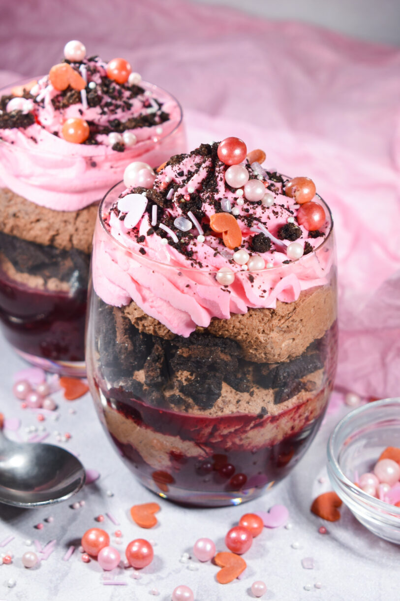 A Valentine's Day dessert parfait in a clear glass showing layers of cherry compote, chocolate mousse, Oreos and pink whipped cream