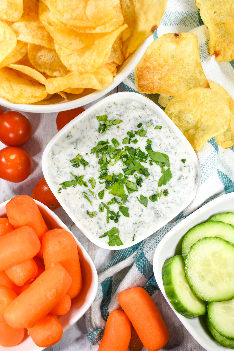 Chip dip with yogurt, alongside potato chips, baby carrots, tomatoes, and cucumbers