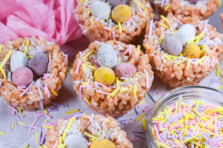 Bird nest rice krispie treats decorated with mini eggs, next to a bowl of pastel sprinkles