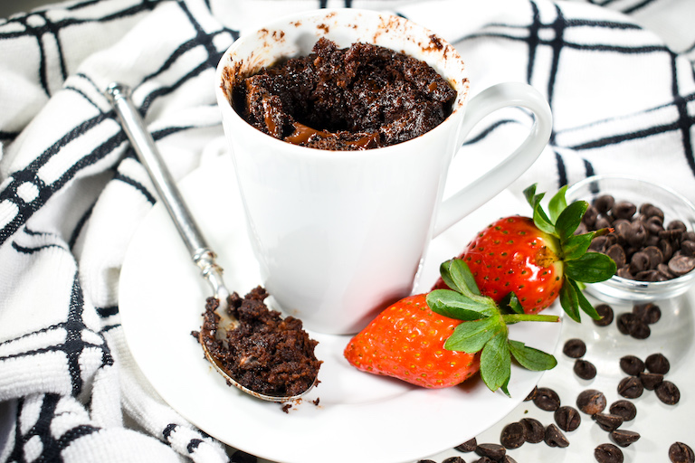 White mug containing a chocolate mug cake, on a plate with a spoon, strawberries, and a bowl of chocolate chips