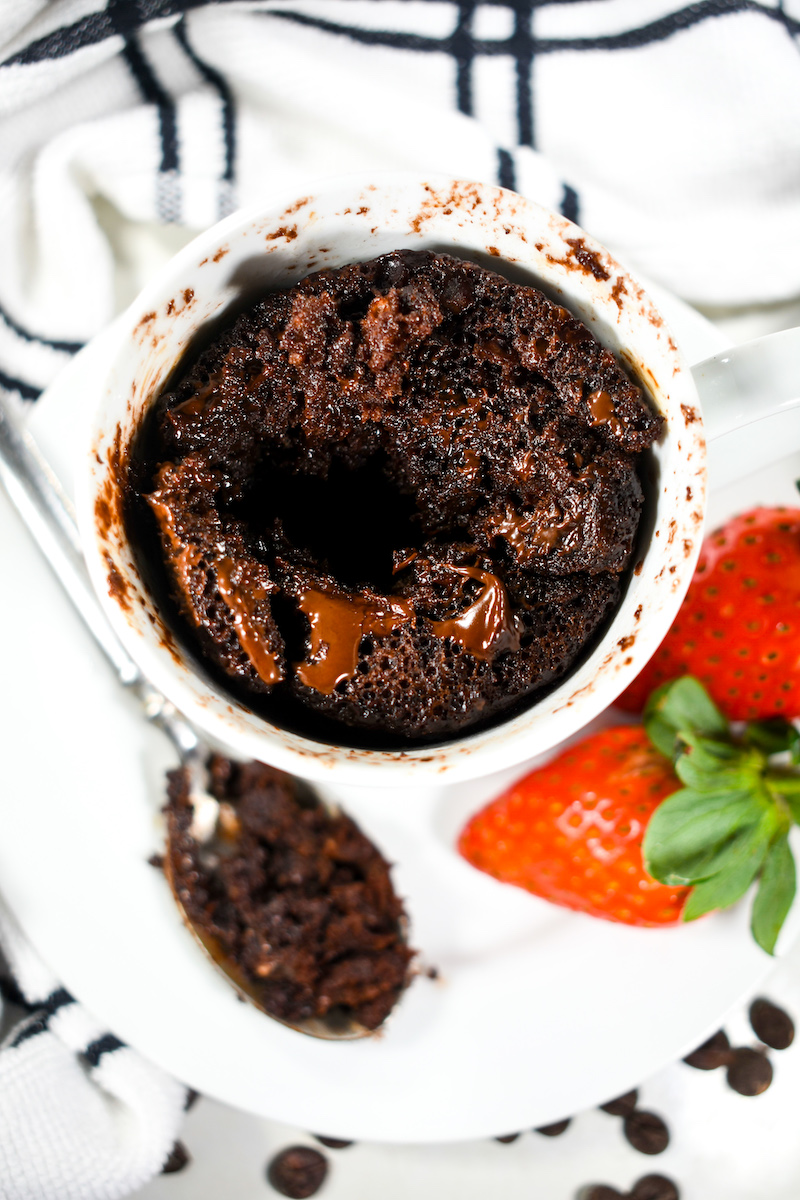 Chocolate cake in a mug, sitting on a plate with a spoon and a pair of red strawberries