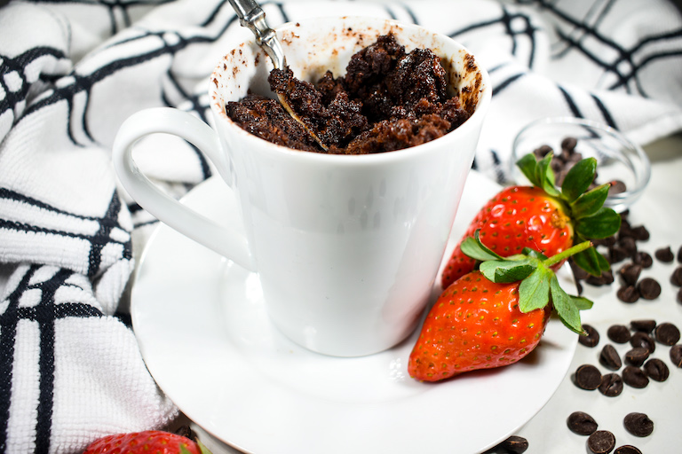 A white mug containing a chocolate mug cake, sitting on a plate with red strawberries