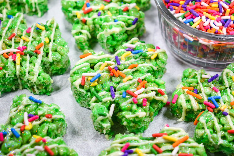 Shamrock rice krispie treats arranged on a white surface with a bowl of rainbow sprinkles