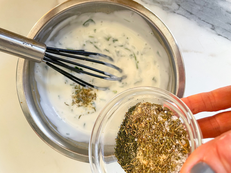 Adding dried herbs and spices to a metal bowl of yogurt dip for veggies