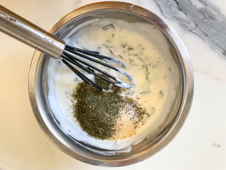 Dried herbs and spices and a whisk in a metal bowl of healthy Greek yogurt dip