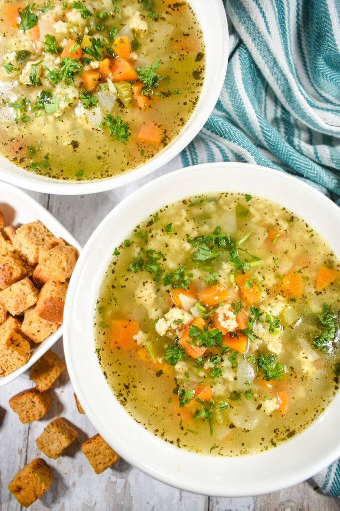 bowls of vegetarian chicken noodle soup on a white surface with a striped towel and dish of croutons