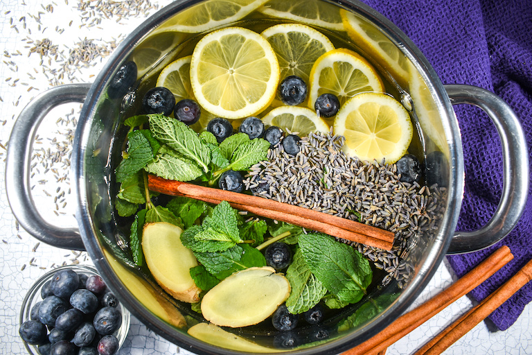 A stock pot filled with cinnamon, mint, ginger, lemon slices, blueberries, and lavender, on a white surface with cinnamon sticks and blueberries