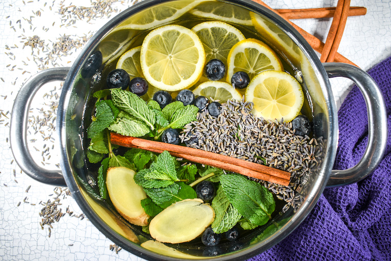 A simmer pot with cinnamon, ginger, mint, blueberries, and lavender, on a white surface with cinnamon sticks and purple towel