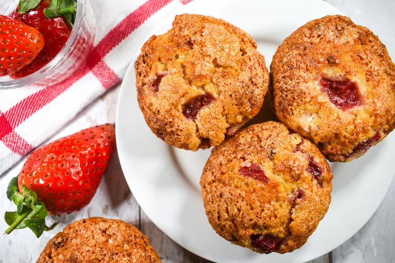 Strawberry muffins on a white plate with a bowl of strawberries and a red and white towel