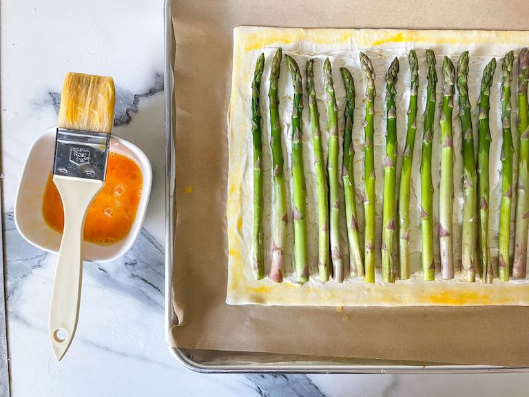 Dish of egg wash, pastry brush, and a rectangle of puff pastry with asparagus spears