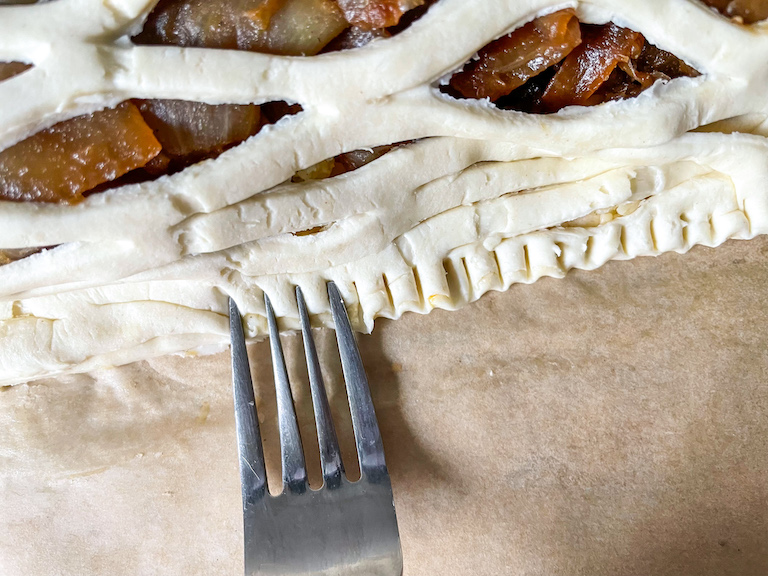 Fork crimping the edges of a lattice puff pastry with cheese and onions