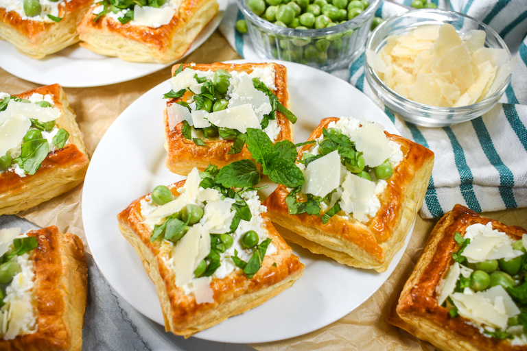 Horizontal photo of puff pastry savory tartlets on a white plate, next to a bowl of peas and bowl of cheese