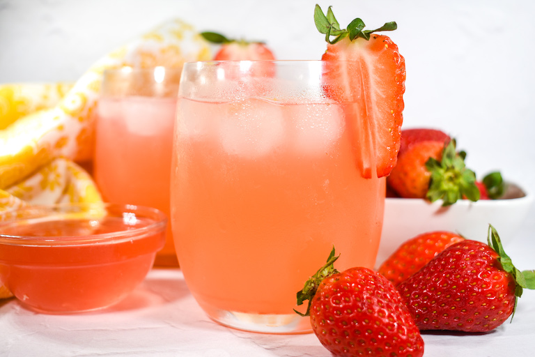 Two glasses of rhubarb and strawwberry lemonade with a dish of simple syrup and fresh strawberries