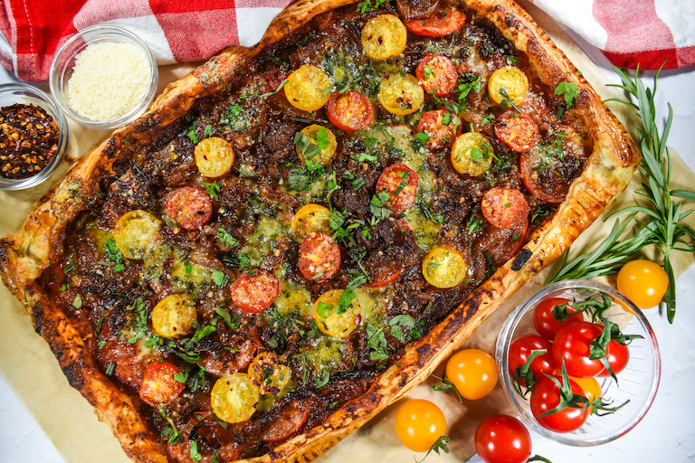 A rectangular shaped cherry tomato tart surrounded by dishes of cheese, pepper flakes, and cherry tomatoes