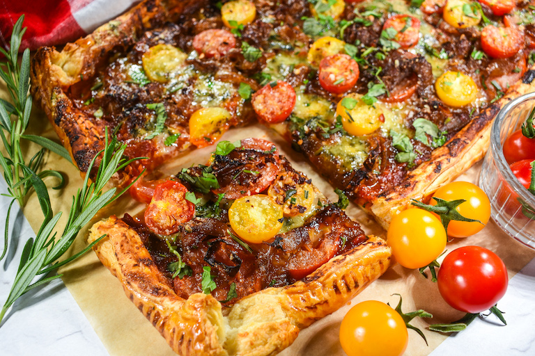 Horizontal shot of a puff pastry tomato galette, cherry tomatoes, and rosemary sprigs on a sheet of parchment