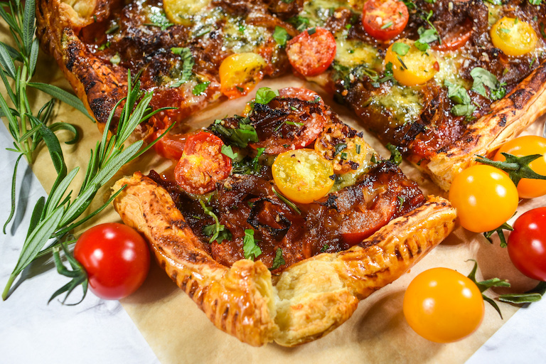 A square slice of cheese and tomato tart surrounded by cherry tomatoes and rosemary