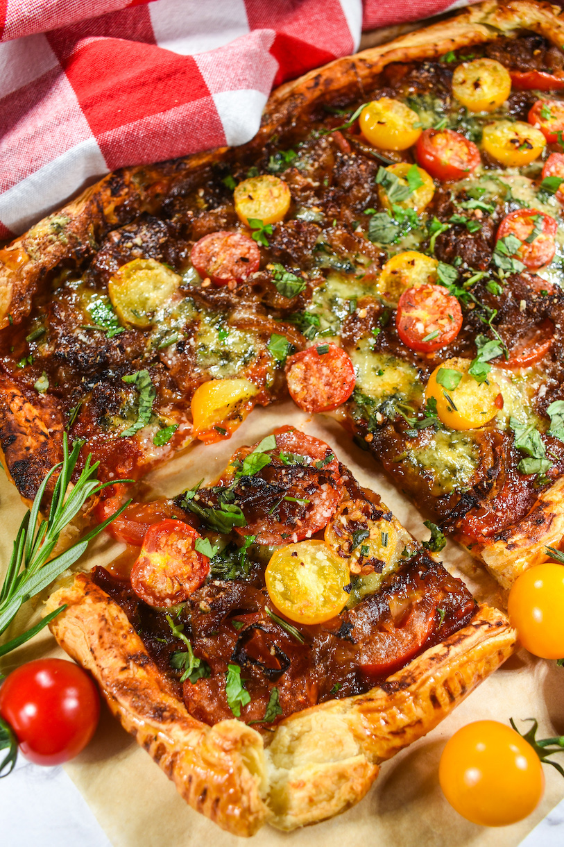 A sliced puff pastry tomato tart surrounded by a red and white towel, cherry tomatoes, and herbs