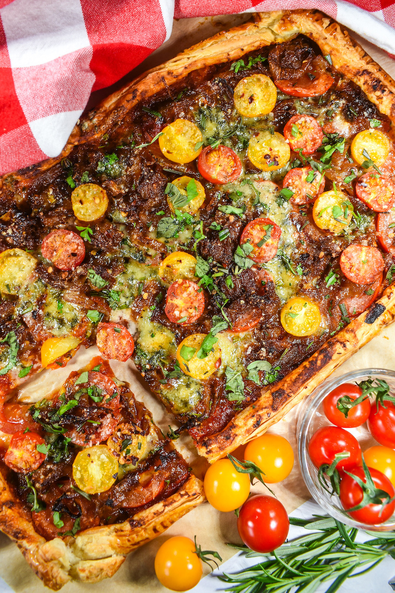 Rustic tomato tart with a square slice removed, alongside a dish fo cherry tomatoes and a sprig of fresh rosemary