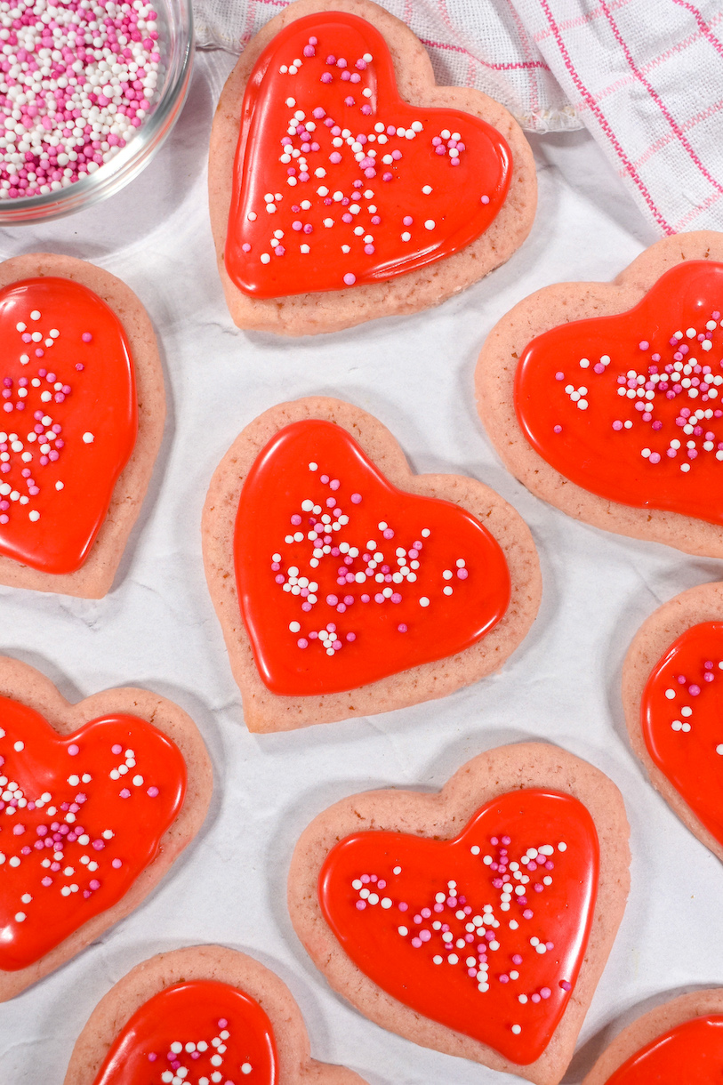 Valentine's heart cookies made from an original Valentine's Day cookie recipe, arranged on a white surface with towel and bowl of sprinkles