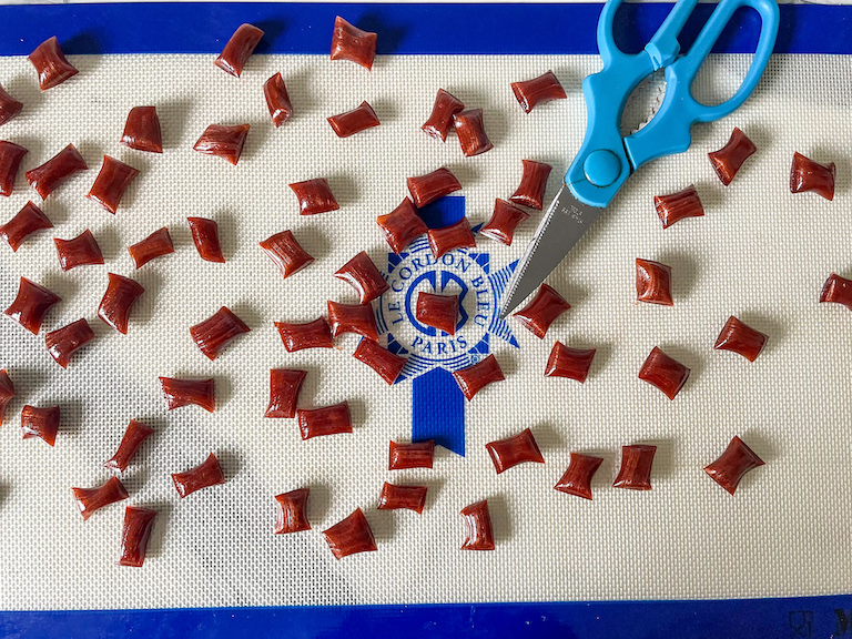 Scissors with blue handles and pieces of homemade root beer hard candy on a silicone mat
