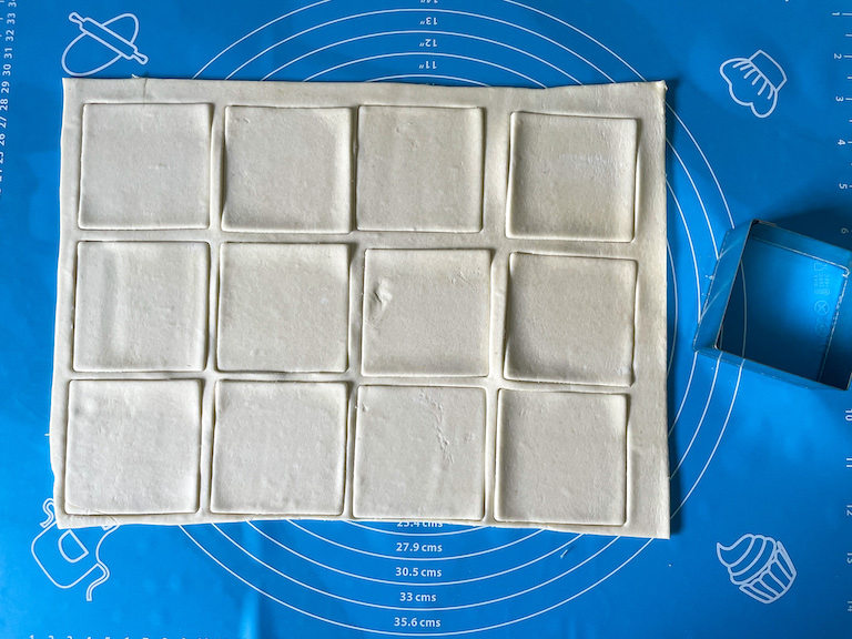 Puff pastry cut into squares and a square metal cutter on a rolling mat