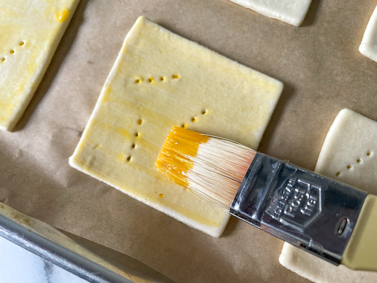 A pastry brush applying egg wash to a square of puff pastry