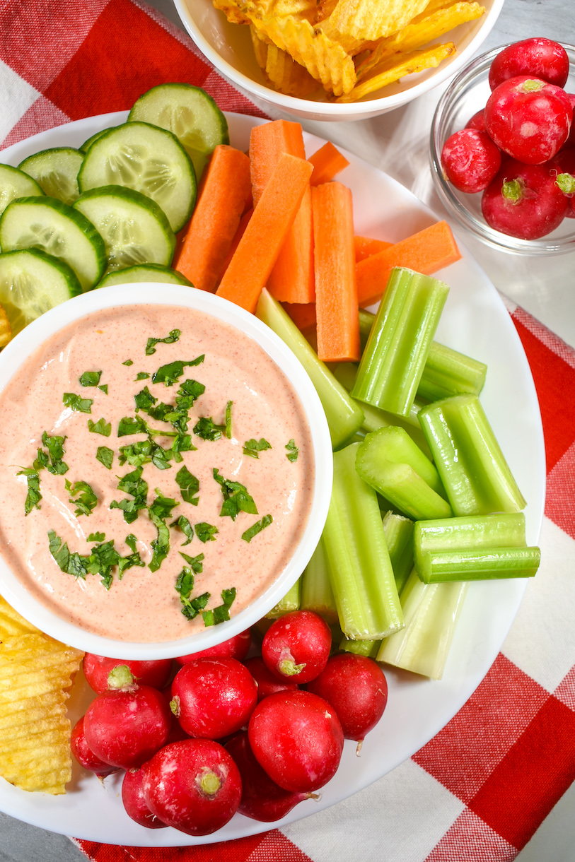 A bowl of sriracha veggie dip with greek yogurt, on a plate surrounded by fresh sliced vegetables