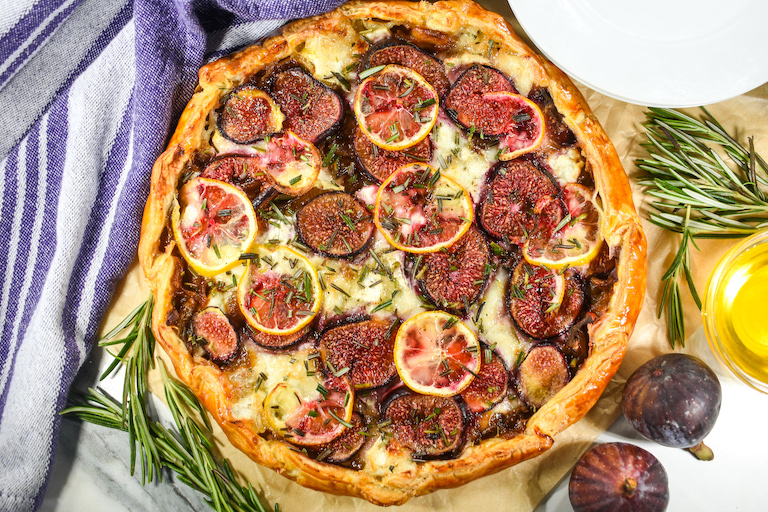 Horizontal shot of a round fig galette arranged on parchment with figs, honey, and rosemary sprigs