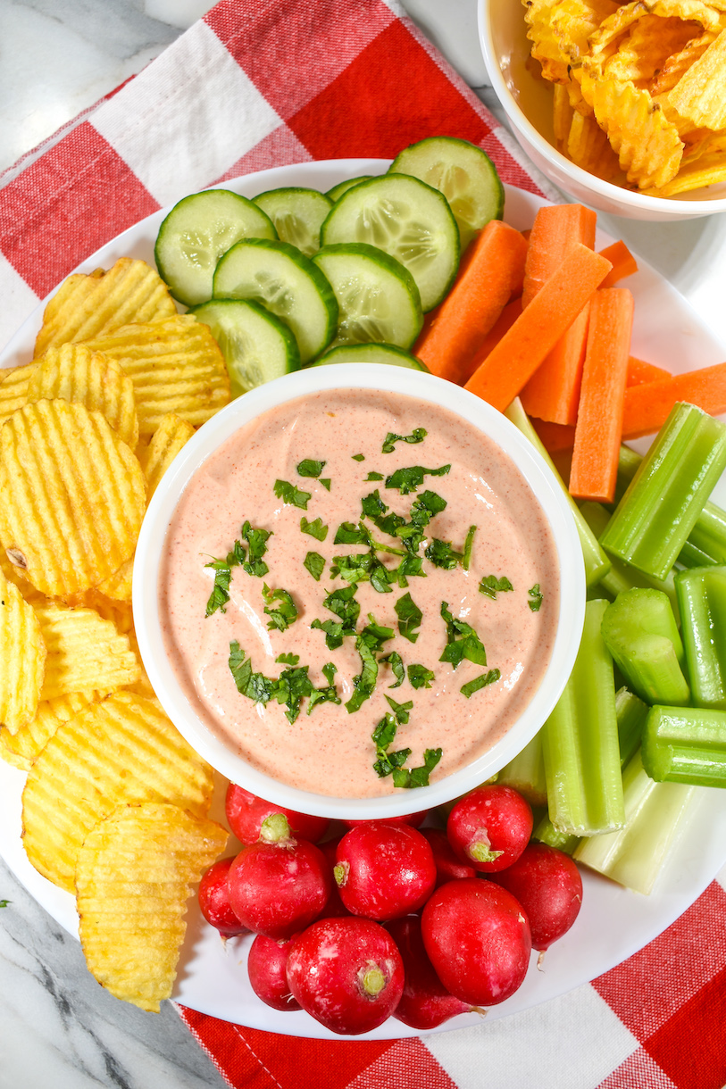 Bowl of sriracha dip on a plate surrounded by chips and raw vegetables