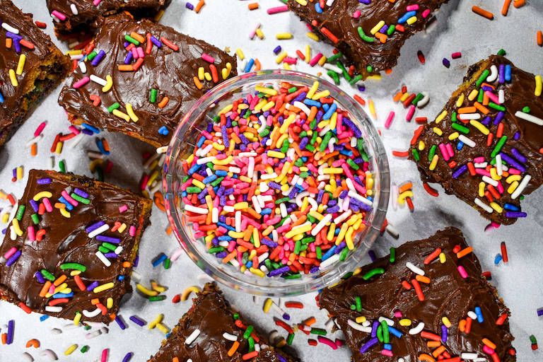 Bowl of sprinkles and frosted chocolate chip cookie bars on a white surface