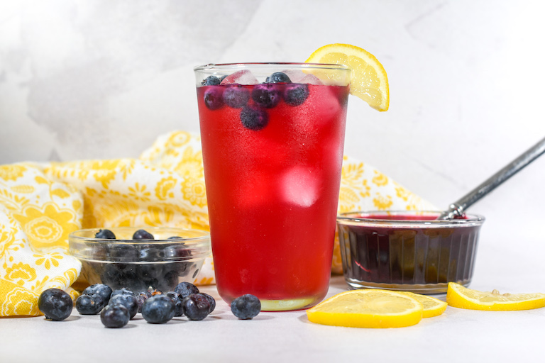 Glass of blueberry lemonade, dish of syrup, fresh berries, and lemon slices