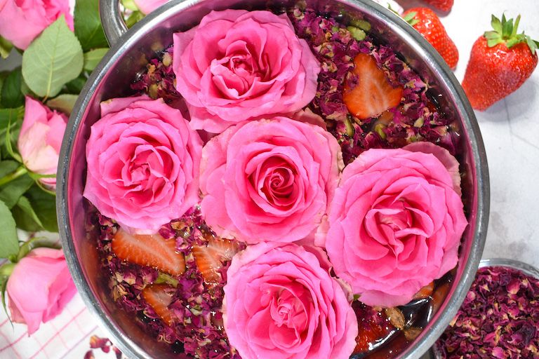 A pot filled with roses and rose petals, surrounded by a bouquet of fresh roses and whole strawberries