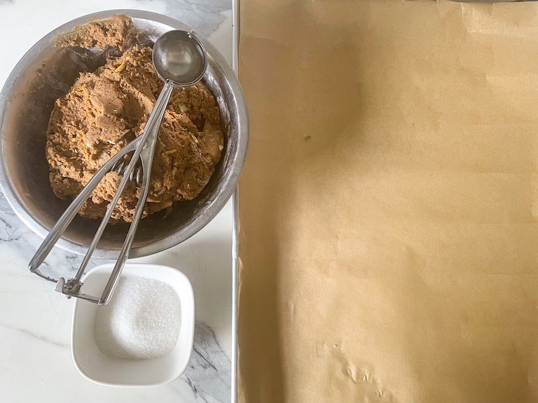 A parchment lined tray, dish of sugar, and bowl of dough with a cookie scoop