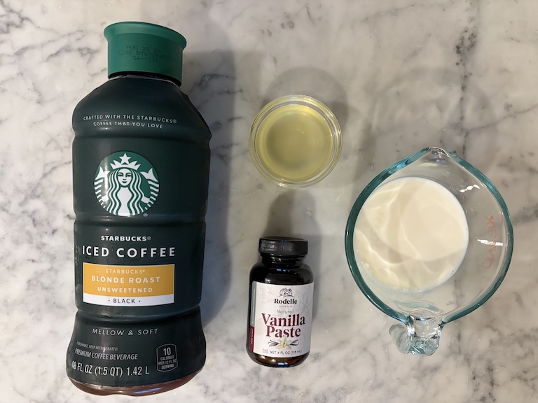 A bottle of iced coffee, jar of vanilla bean paste, dish of orange syrup, and cup of milk arranged on a marble surface