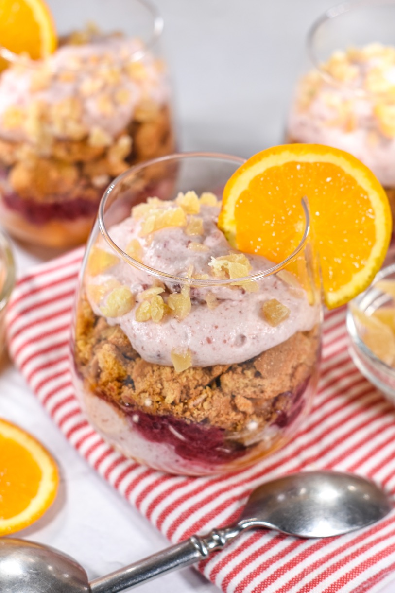 Cranberry and orange parfait in a clear wine glass, garnished with a slice of fresh orange, with a striped towel and a spoon