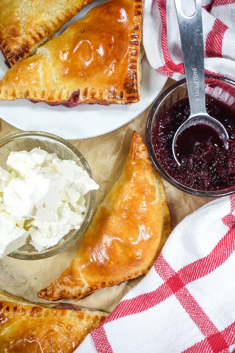 Looking down at puff pastry triangles, a red and white towel, and dishes of cheese and cherries