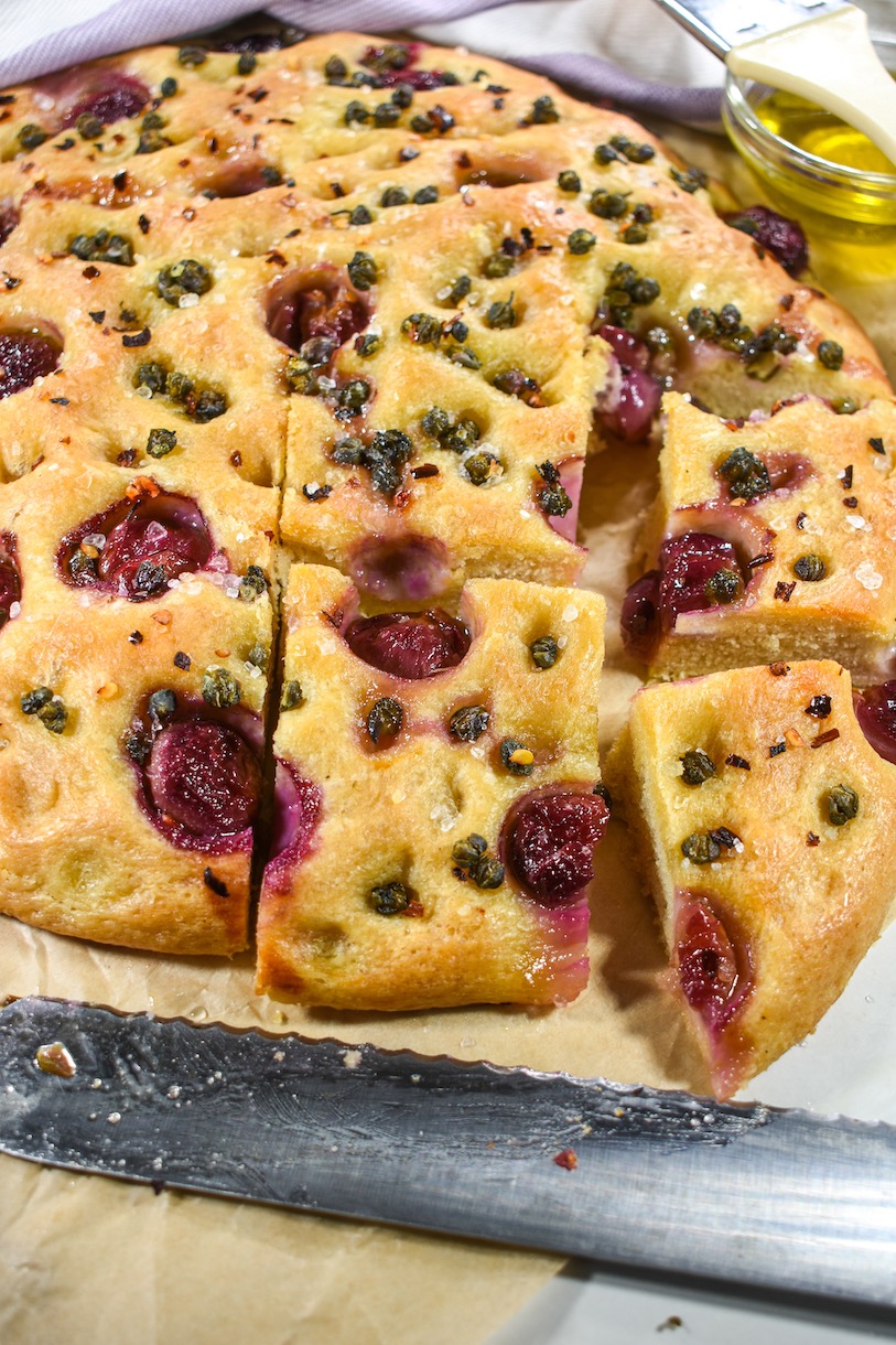 A sliced loaf of focaccia bread with grapes and capers