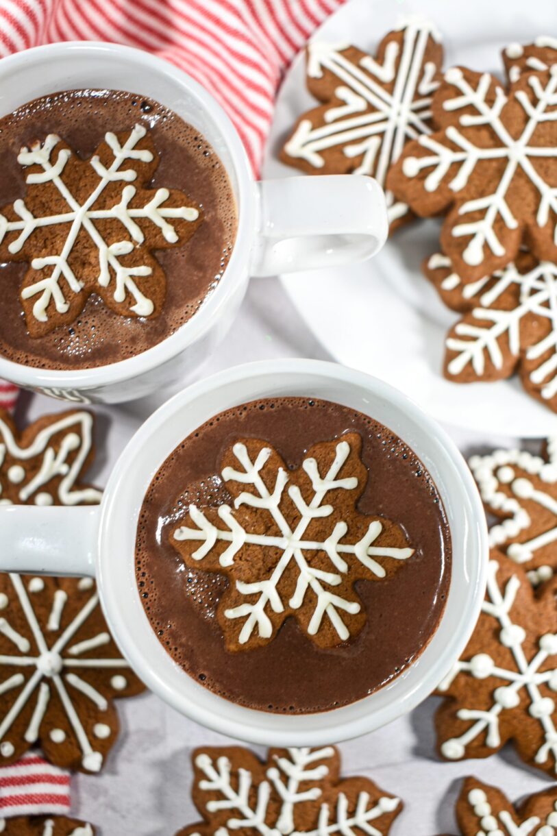 Mugs of gingerbread hot chocolate surrounded by snowflake shaped gingerbread cookies and a striped towel