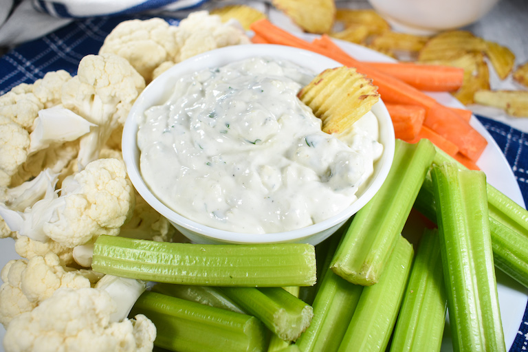 Horizontal image of a bowl of blue cheese dip surrounded by celery sticks and raw cauliflower florets