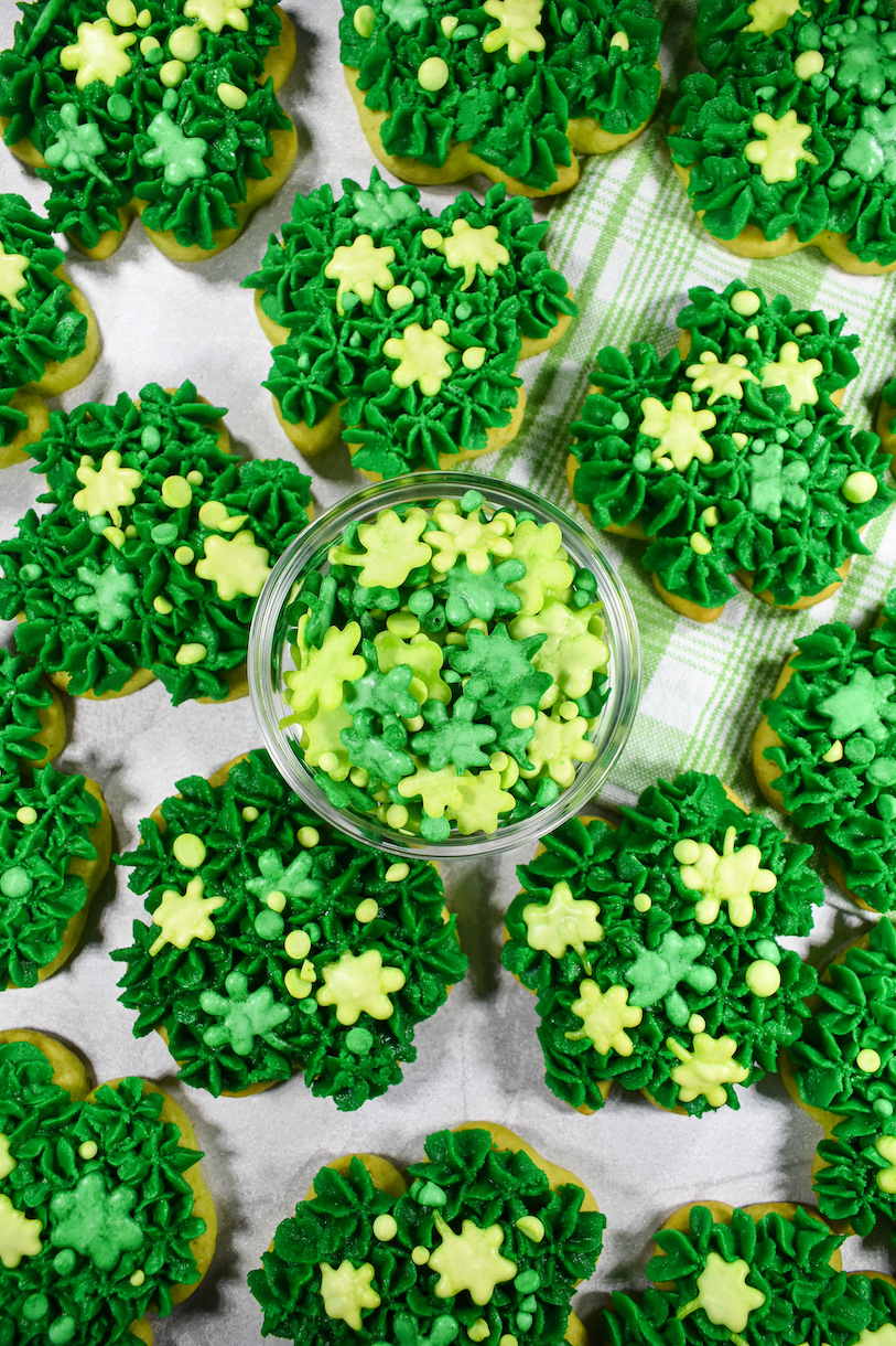 Buttercream frosted sugar cookies shaped like shamrocks, surrounding a bowl of shamrock sprinkles