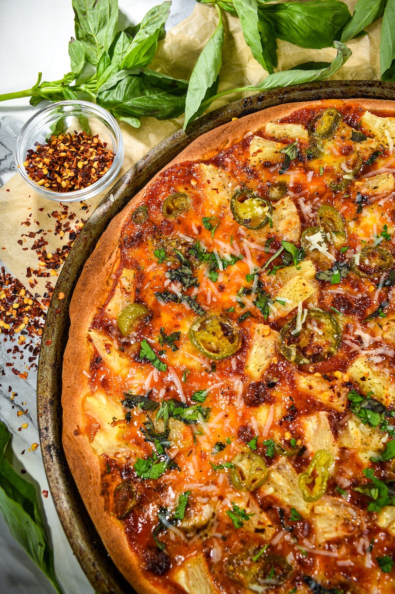 Pineapple pizza with a bowl of chili flakes, and a sprig of fresh basil