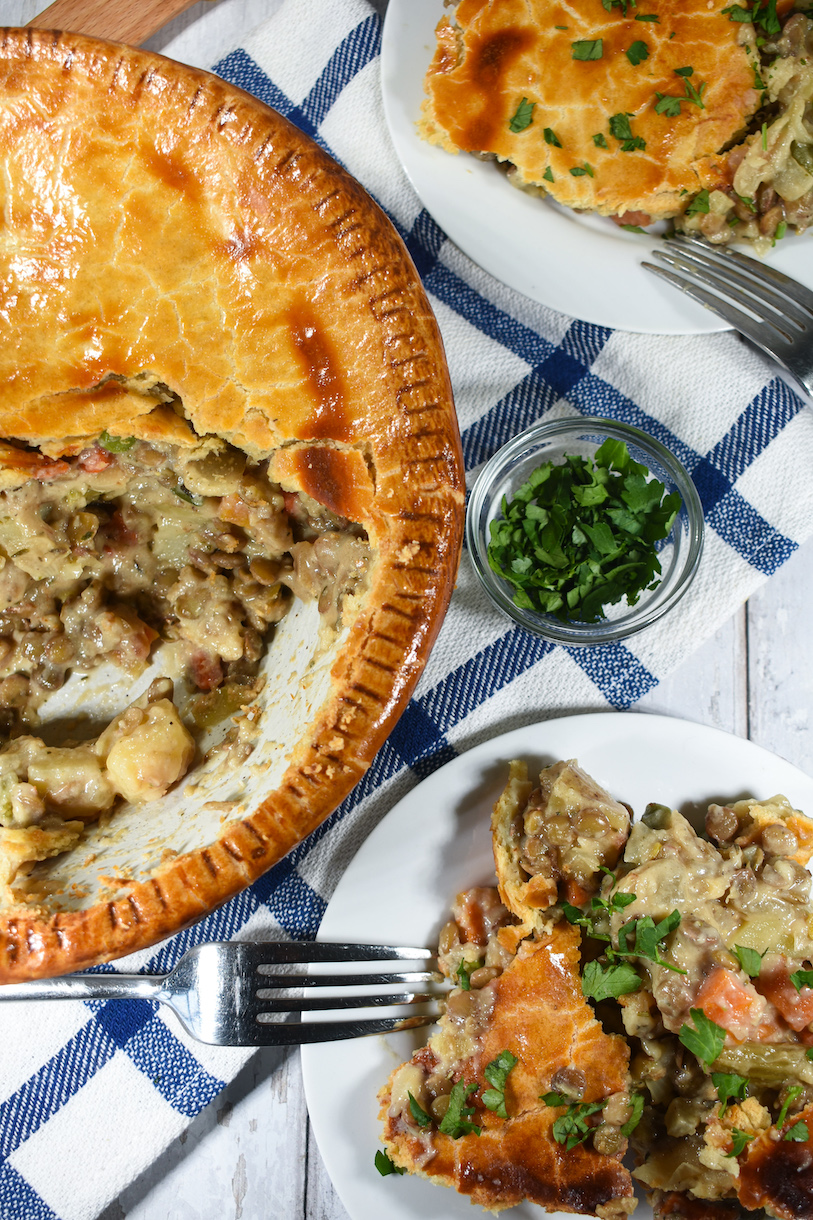 Vegetarian pot pie, blue plaid tea towel, bowl of herbs, and a fork on a white surface