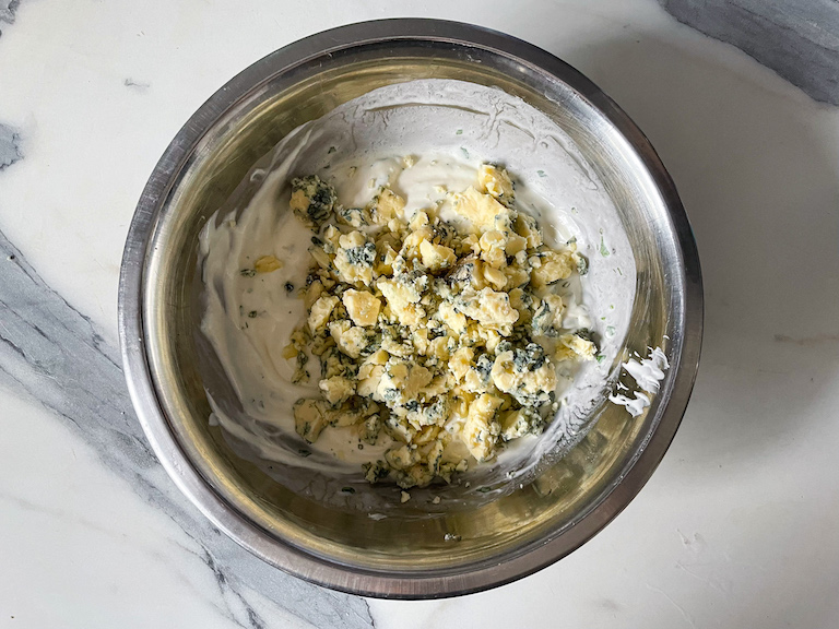 Adding crumbled blue cheese to a bowl of mayonnaise and sour cream